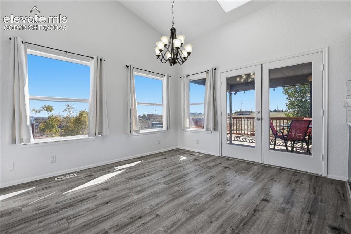 Vaulted ceiling with a multi-arm chandelier, wood-finish flooring, three large picture windows, and double glass-paned doors opening to an exterior deck