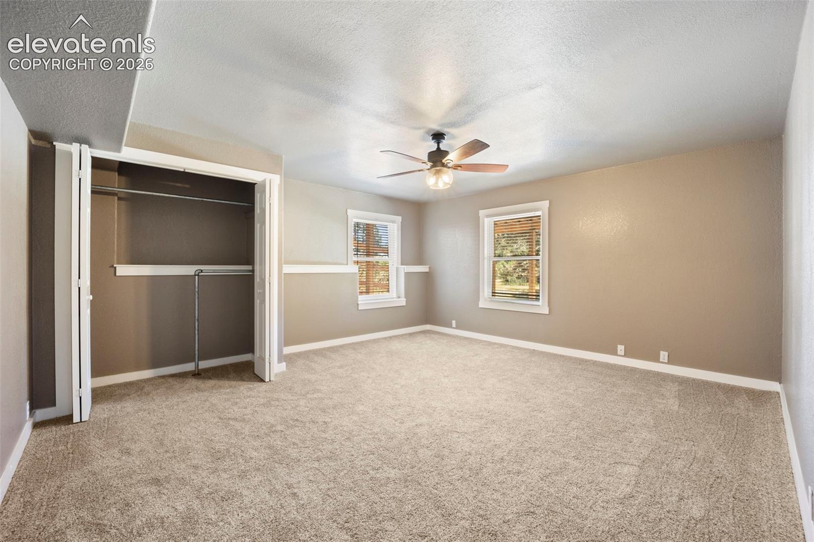 Spacious room featuring neutral carpeting, two windows with white trim, a ceiling fan with light fixture, and a bi-fold closet