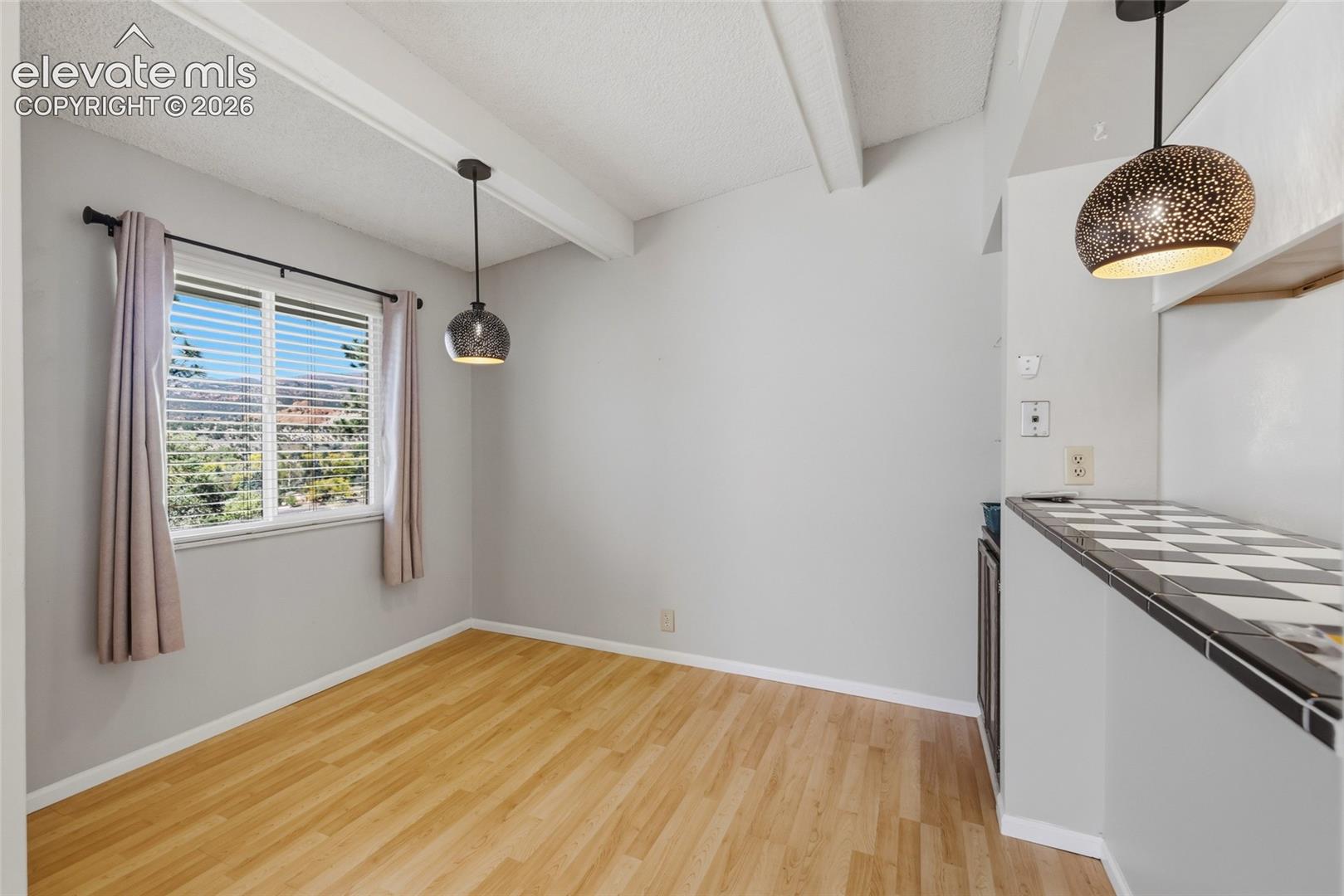 Room featuring wood-finish flooring, vaulted ceiling with exposed beams, two metallic pendant lights, and a counter with a checkerboard tile surface