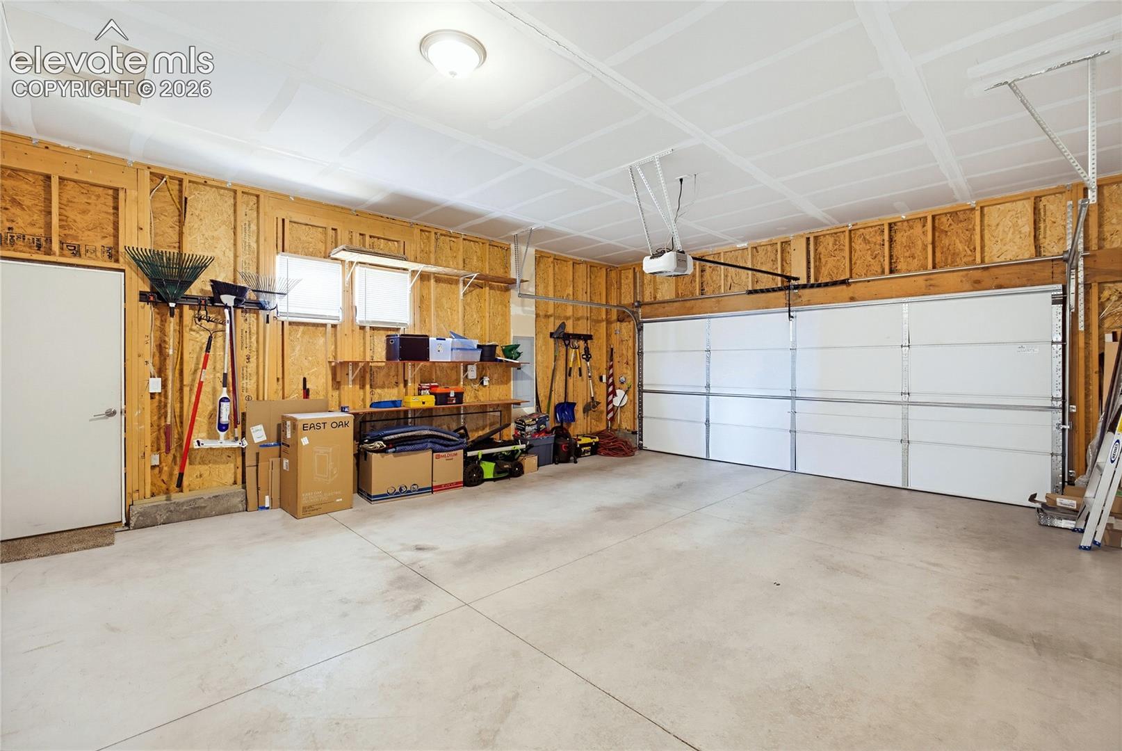 Attached two-car garage featuring a concrete floor, white sectional door, exposed wood-stud walls, built-in shelving, and a dedicated service door