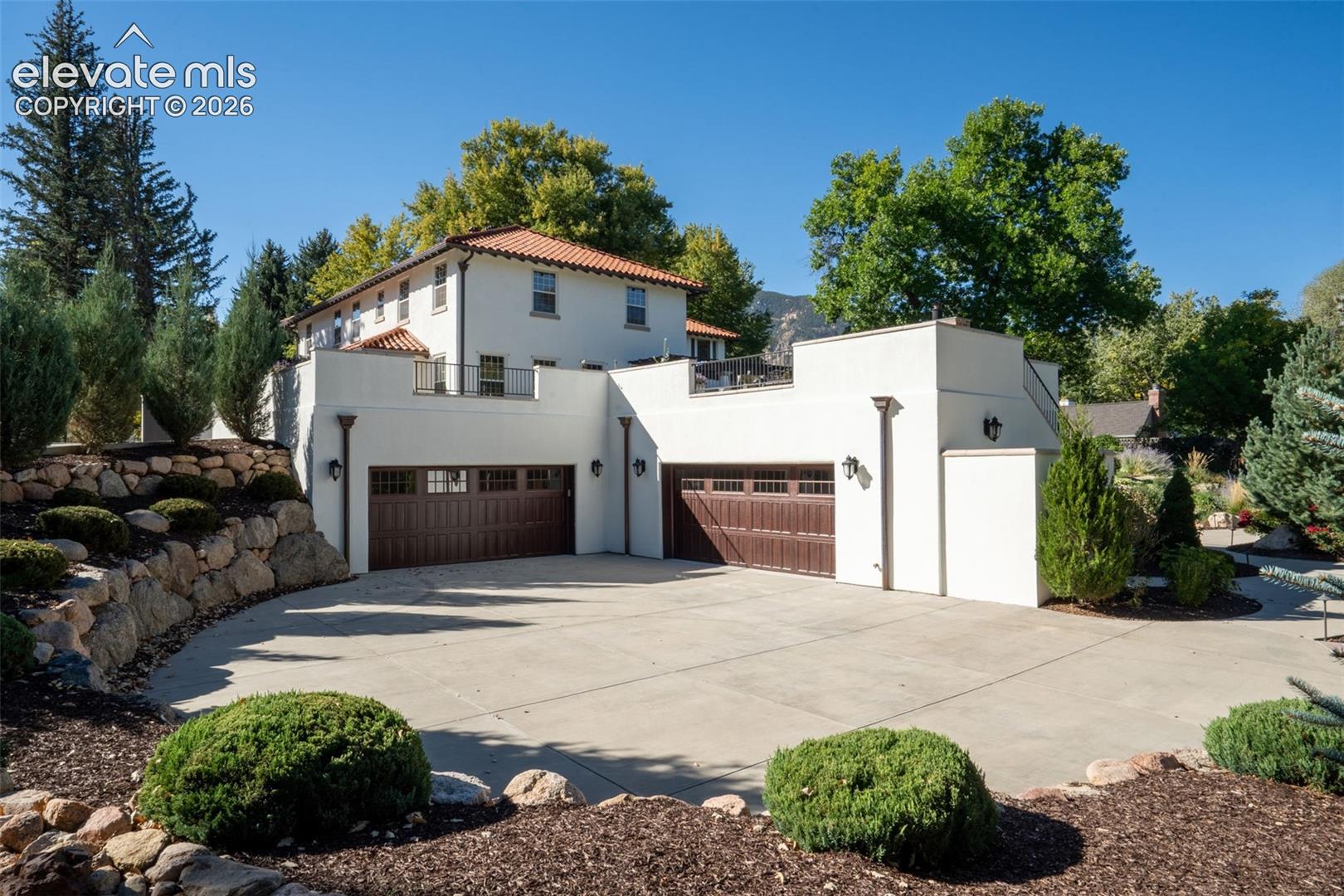 Stucco exterior featuring a terracotta tile roof, two-car garage with paneled doors, expansive upper-level terrace, and stone retaining walls