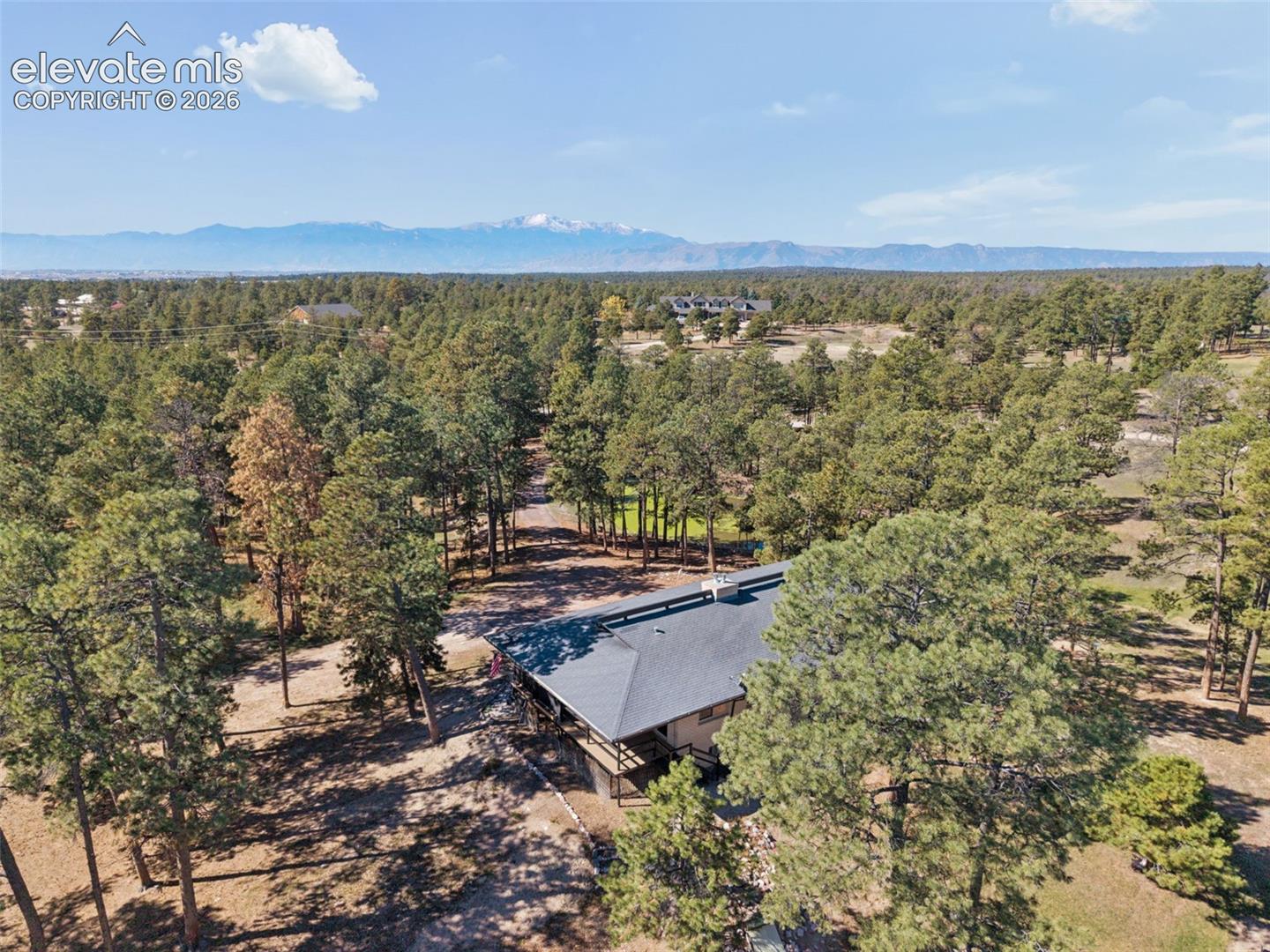 Aerial perspective of a sprawling property featuring mature evergreen trees, a multi-faceted roofline, wrap-around decking, and a prominent mountain range backdrop