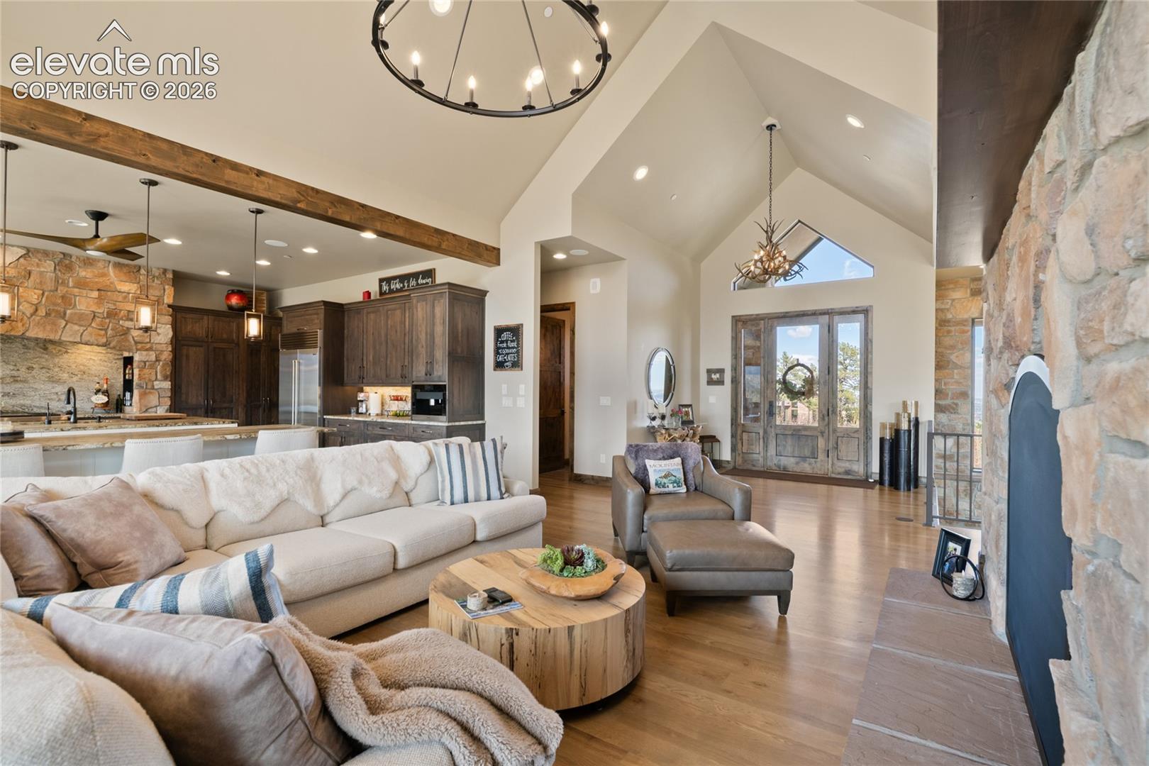 Where the whole home comes together. Open-concept great room showcasing soaring vaulted ceilings, exposed wood beams, statement chandeliers, floor-to-ceiling stone fireplace, and seamless flow from the chef's kitchen to the formal entry.