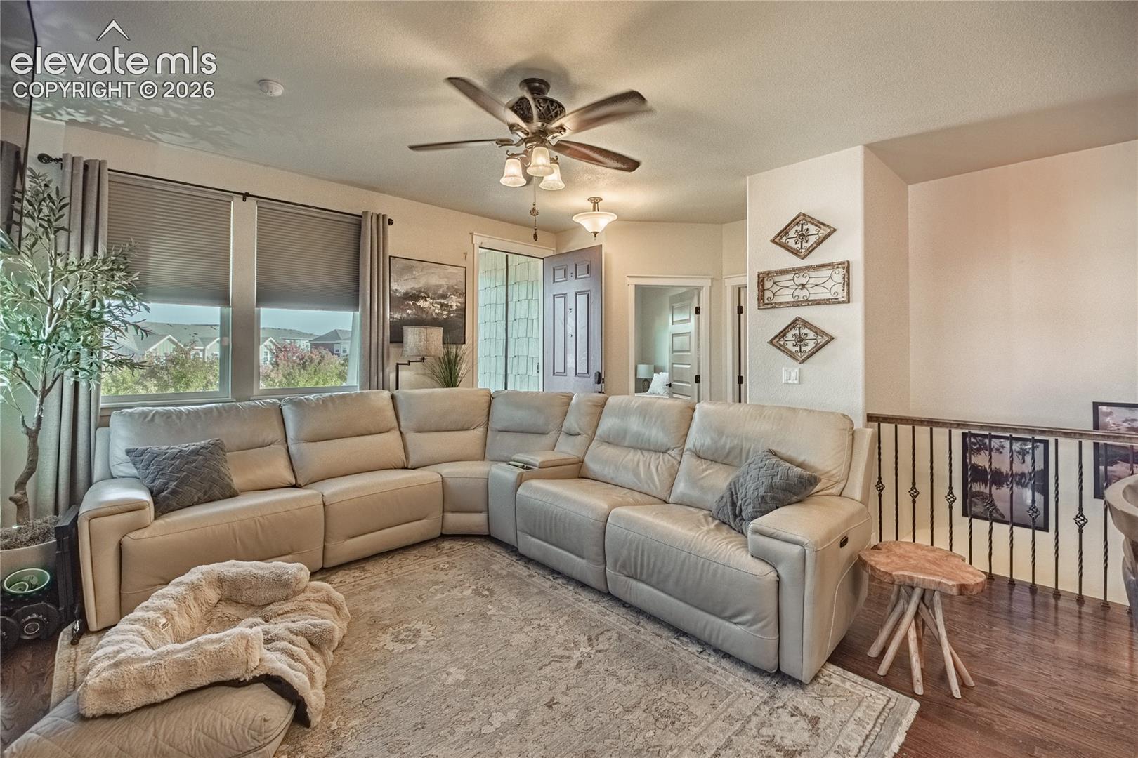Open-concept space featuring wood-finish flooring, a multi-blade ceiling fan with integrated lighting, dual window treatments, and ornate metal stair railings