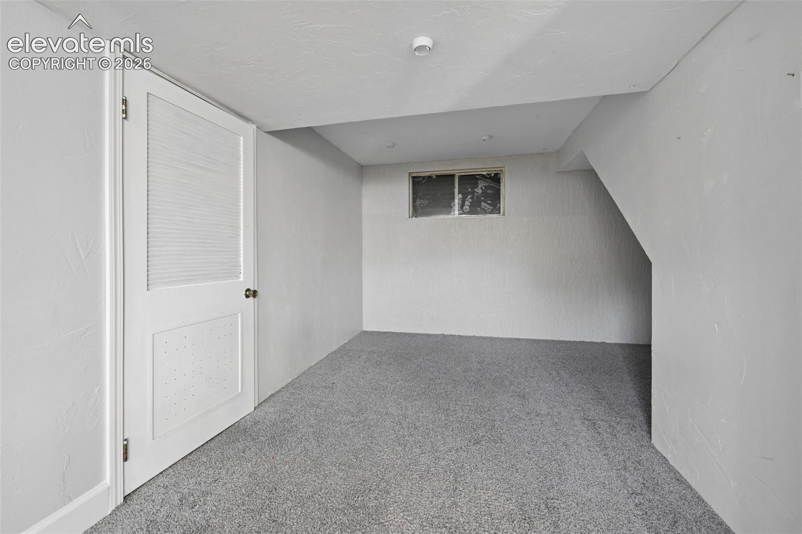 Basement with utility closest and extra “nook” area makes for a great play area!  featuring neutral-toned carpeting, textured walls, a solid-core louvered panel door, and window