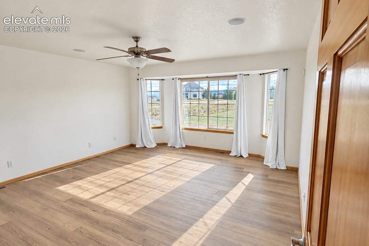 Main-level primary. Spacious room featuring wood-finish flooring, a multi-pane bay window, built-in ceiling speakers, and a ceiling fan with integrated lighting. No staging