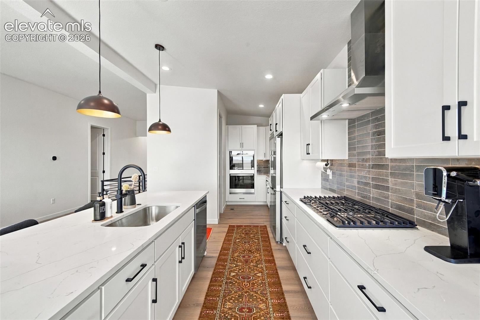 Kitchen featuring white cabinetry with matte black hardware, quartz countertops, a horizontal tile backsplash, stainless steel gas cooktop with chimney-style range hood, and dual pendant lighting