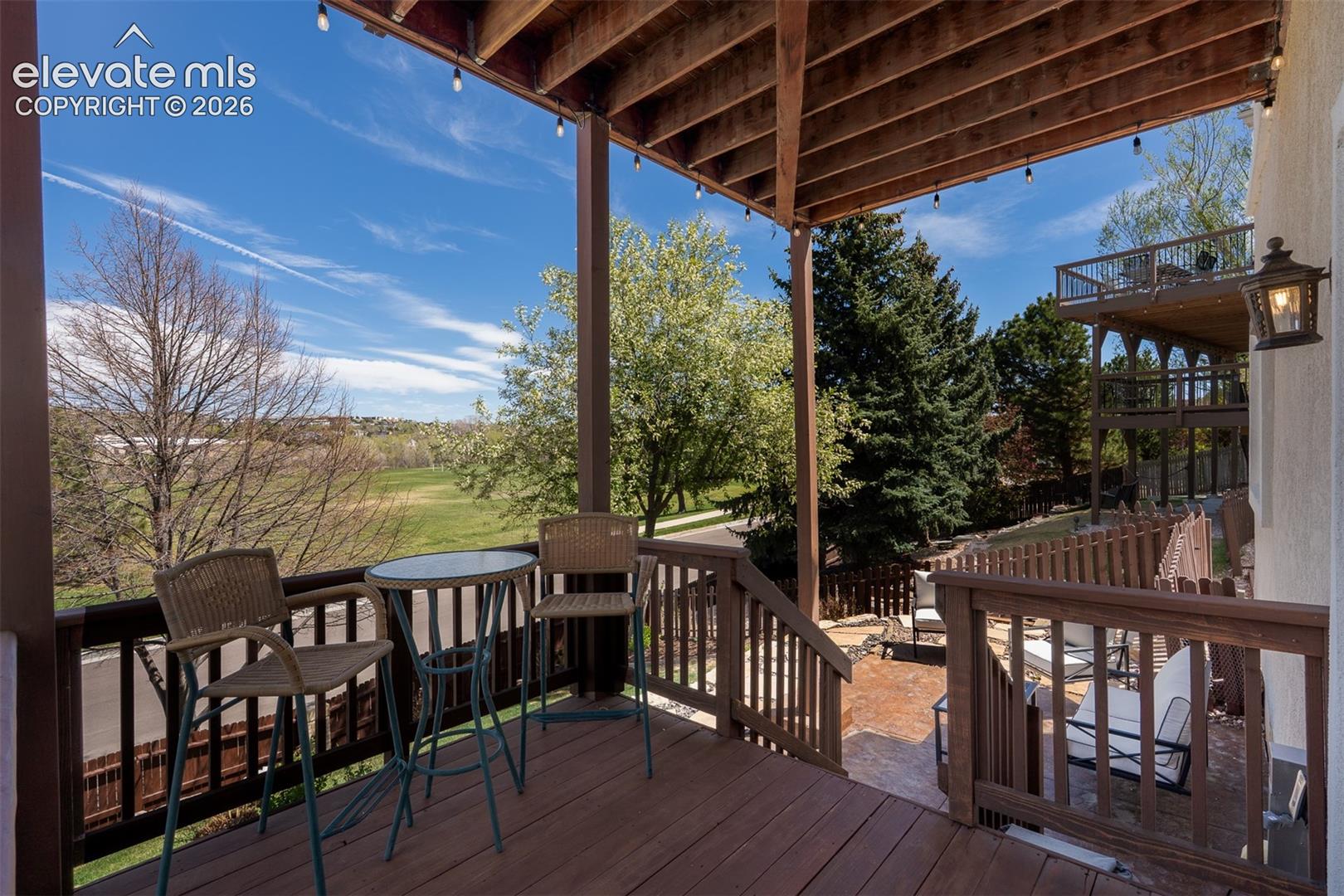 Walk out lower level onto the lower deck featuring dark wood railing, exposed ceiling joists, string lighting, and a terraced backyard with mature trees.  Note the sitting area on decortive concrete pad.