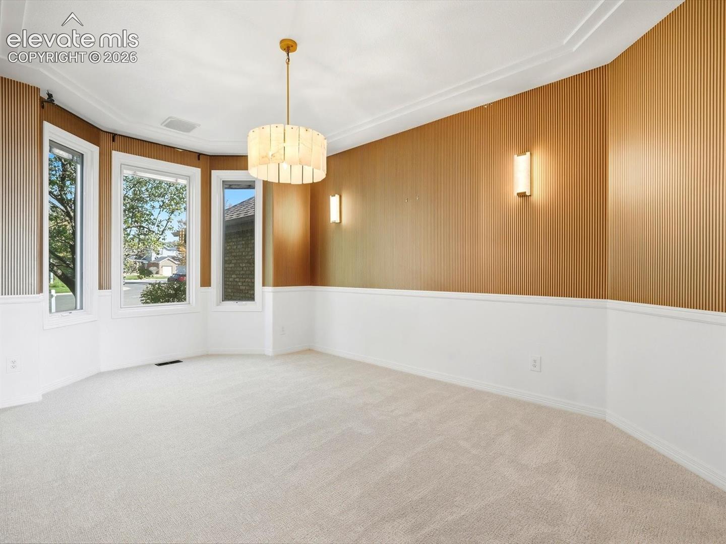 Room featuring bay windows, vertical wood-panelled accent walls, wainscoting, a tiered brass chandelier, and wall-mounted sconces