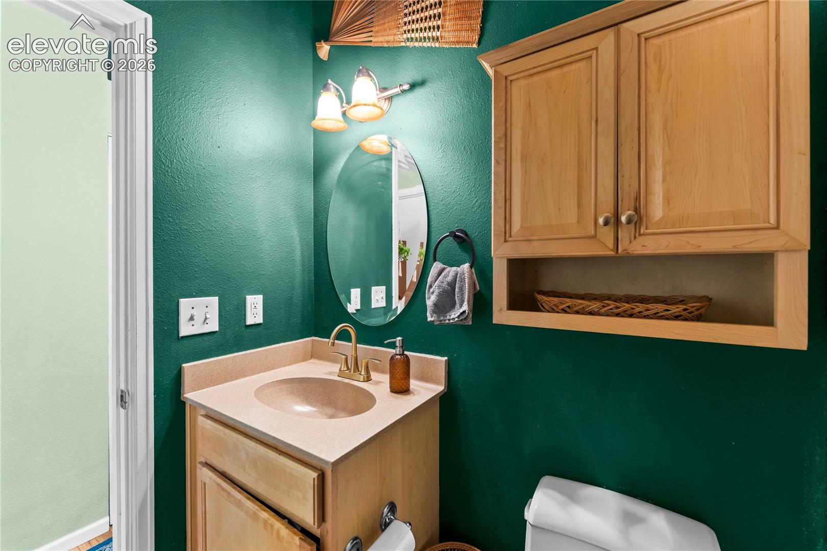 Vanity featuring light wood-finish cabinetry, a solid-surface countertop with an integrated basin, and a brass-tone faucet, complemented by a wall-mounted wood-finish cabinet and an oval mirror