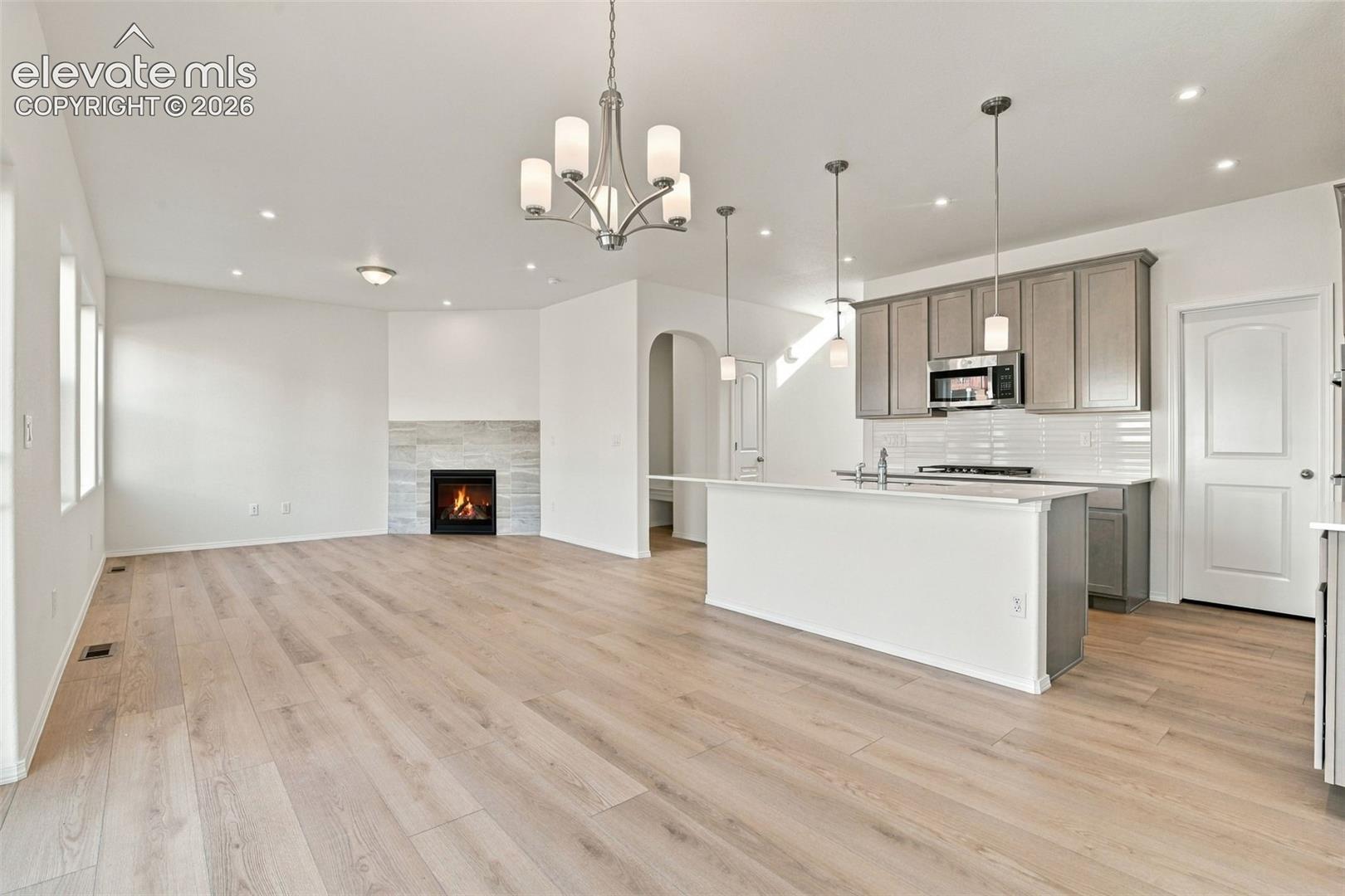 Open-concept kitchen and living area featuring wood-finish flooring, recessed lighting, a stone-surround fireplace, a center kitchen island with pendant lighting, and shaker-style cabinetry