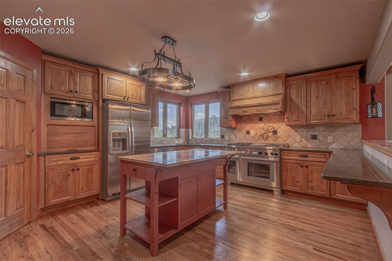 Kitchen featuring custom wood cabinetry, stainless steel appliances, a stone tile backsplash, a center island with built-in shelving, and wood-finish flooring