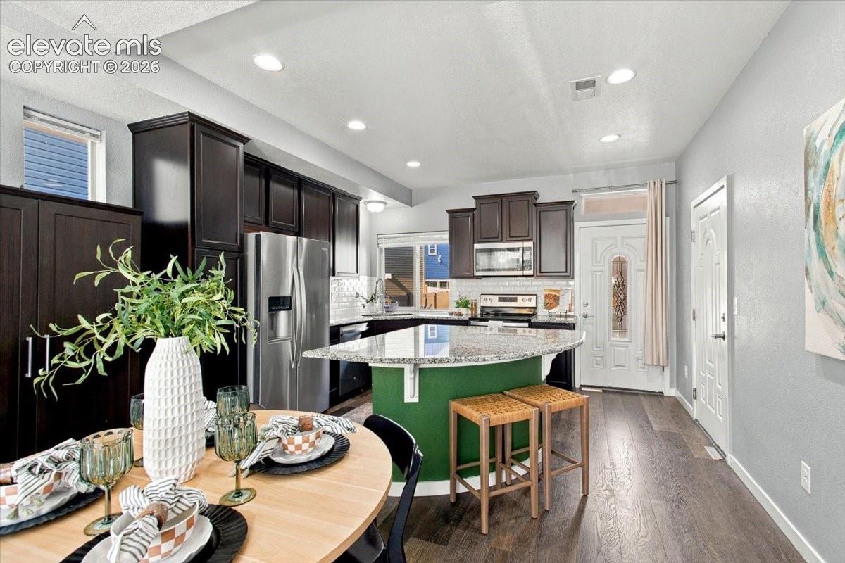 Dark wood cabinetry, stainless steel refrigerator, kitchen island with granite countertop, wood-finish flooring, and recessed lighting