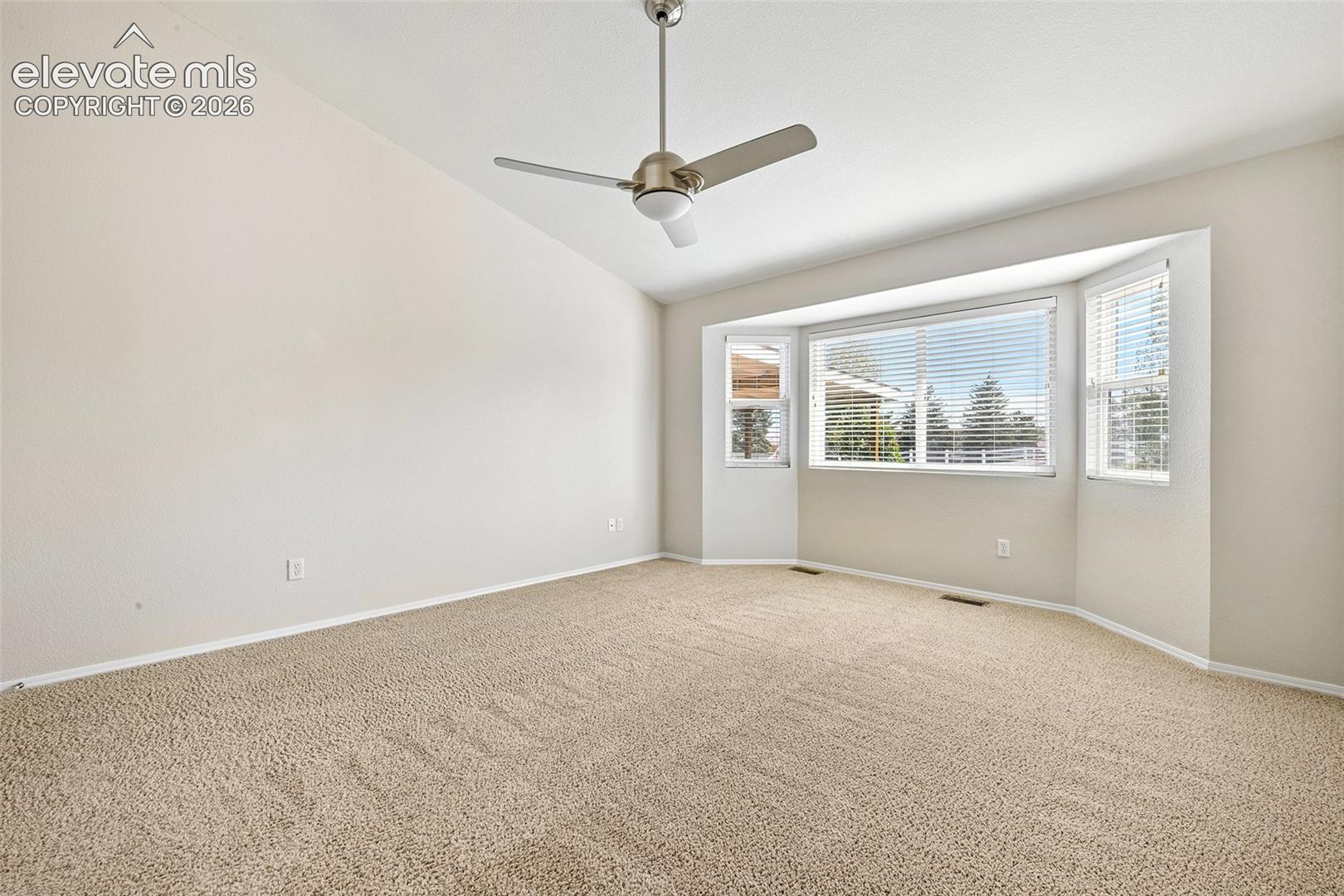 Vaulted ceiling, bay window, light-colored carpeting, and a ceiling fan