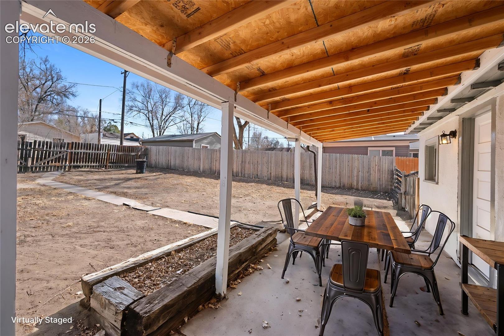 Covered patio featuring an exposed timber-beam ceiling, concrete flooring, and a timber-retained raised planter box, complemented by a fenced backyard with a concrete pathway