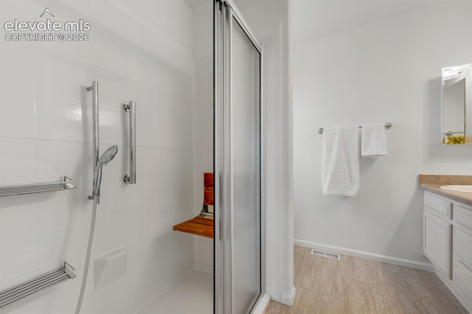 Walk-in shower featuring a glass enclosure, integrated wooden bench, handheld shower head, and dual metal shelving units, paired with a white vanity and wood-finish flooring