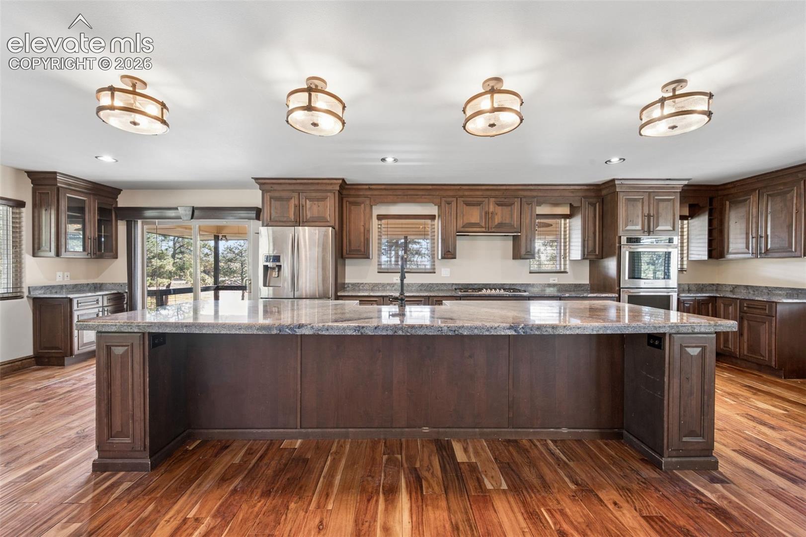 Spacious kitchen featuring an expansive center island, custom wood cabinetry, stone countertops, wood-finish flooring, and built-in stainless steel appliances