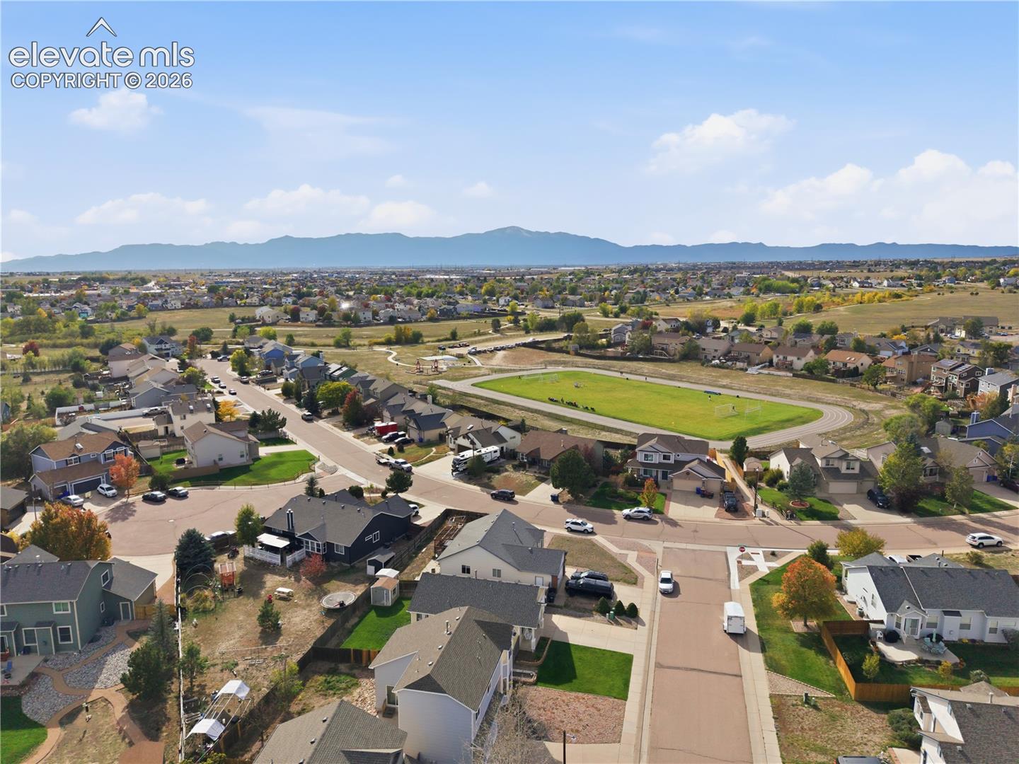 Residential neighborhood with diverse home styles, paved streets, and a prominent green field, all framed by distant mountains