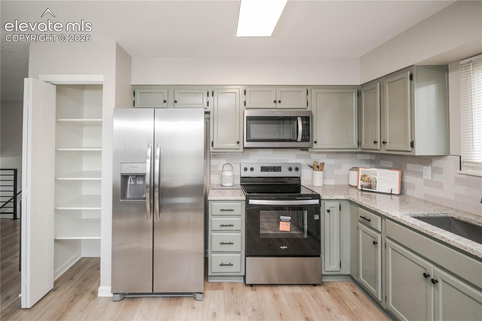 Kitchen featuring sage cabinetry, light-toned stone-finish countertops, stainless steel appliances, a tiled backsplash, and a dedicated pantry with built-in shelving
