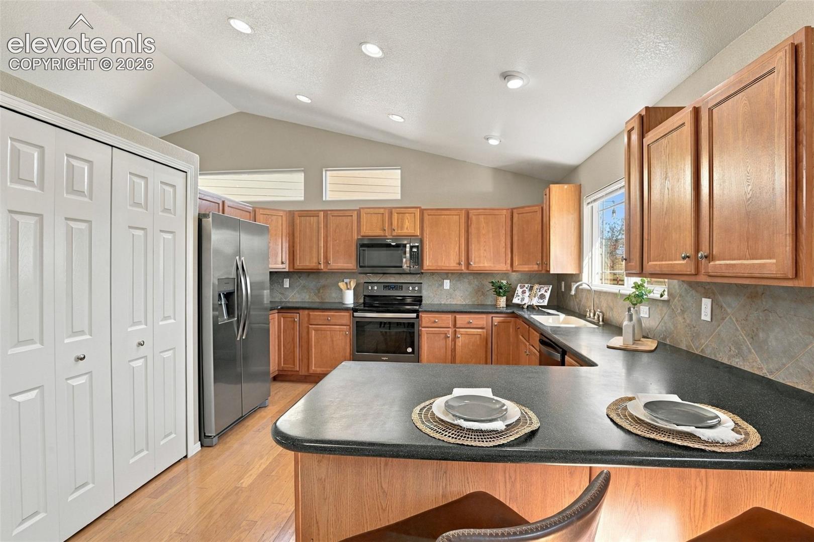 Kitchen featuring wood cabinetry, stainless steel appliances, dark countertops, a tile backsplash, and vaulted ceilings with recessed lighting