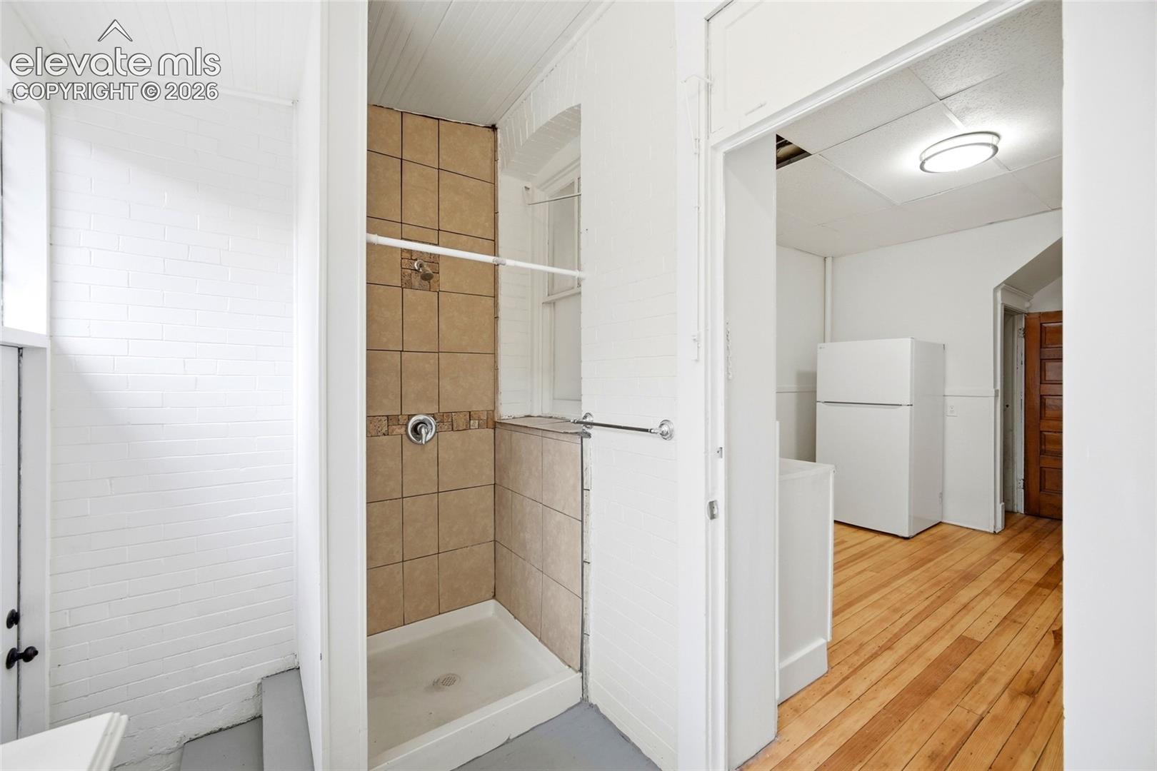 Shower stall featuring tiled surround and metallic fixtures, complemented by wood-finish flooring and white-painted brick interior walls