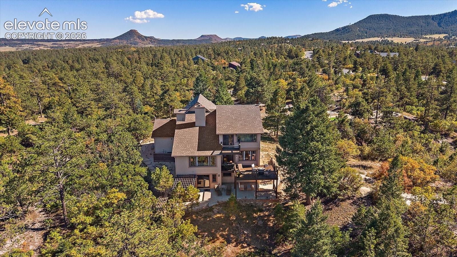 Multi-story residence featuring a shingled roof, attached elevated deck, rear stone patio, and extensive surrounding woodland with a mountain backdrop
