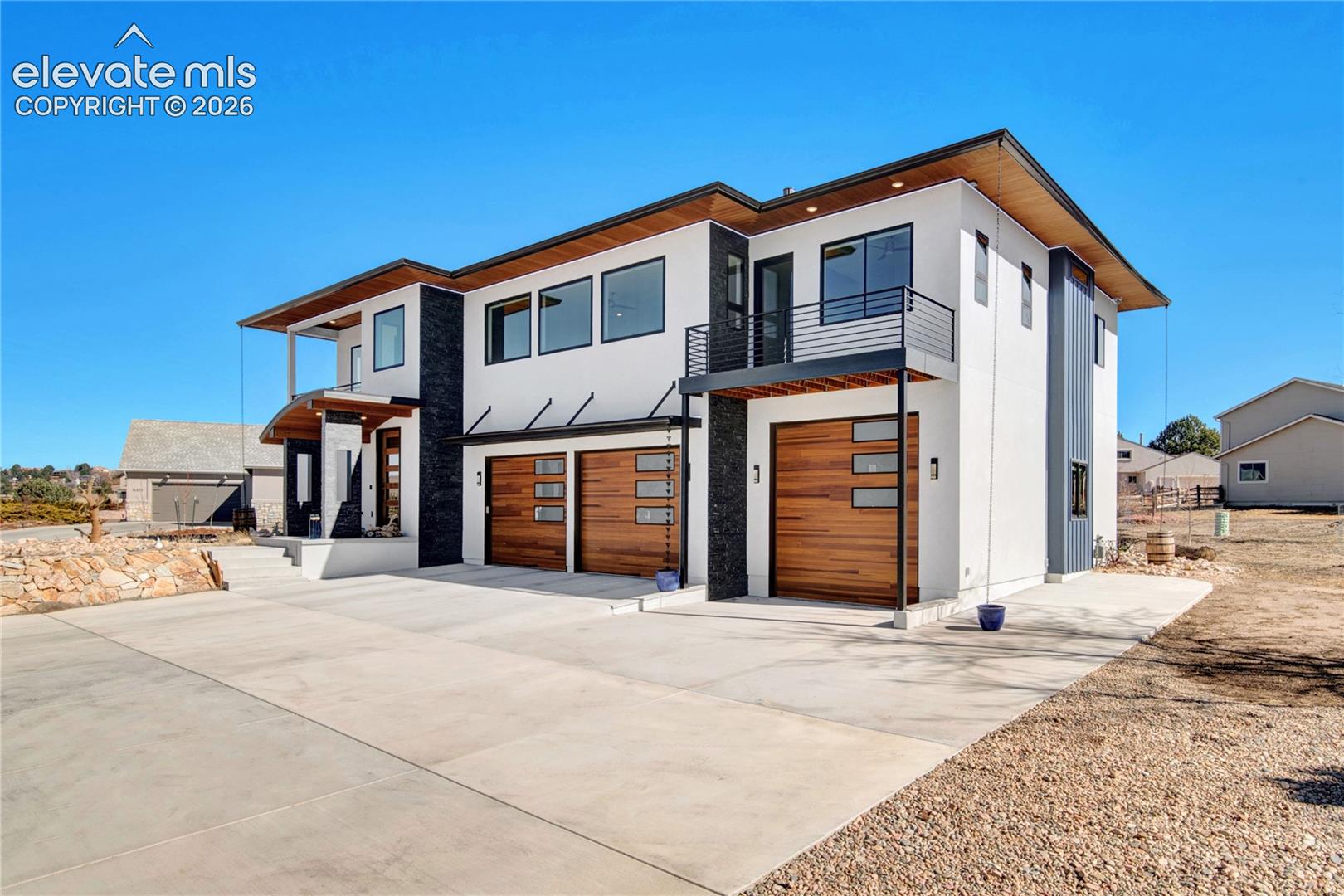Modern architectural design featuring a white stucco exterior with dark accent panels, wood-finish garage doors, and multiple balconies