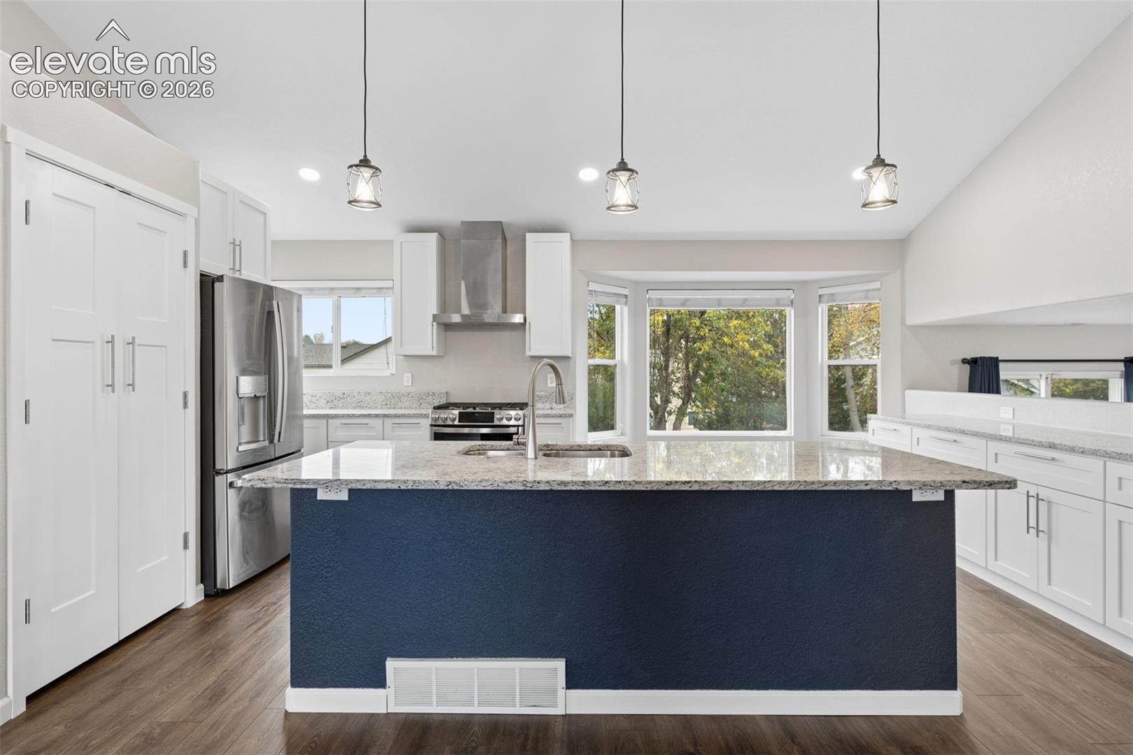 Kitchen featuring a large central island with a stone-finish countertop, white shaker-style cabinetry, stainless steel appliances, pendant lighting, and wood-finish flooring