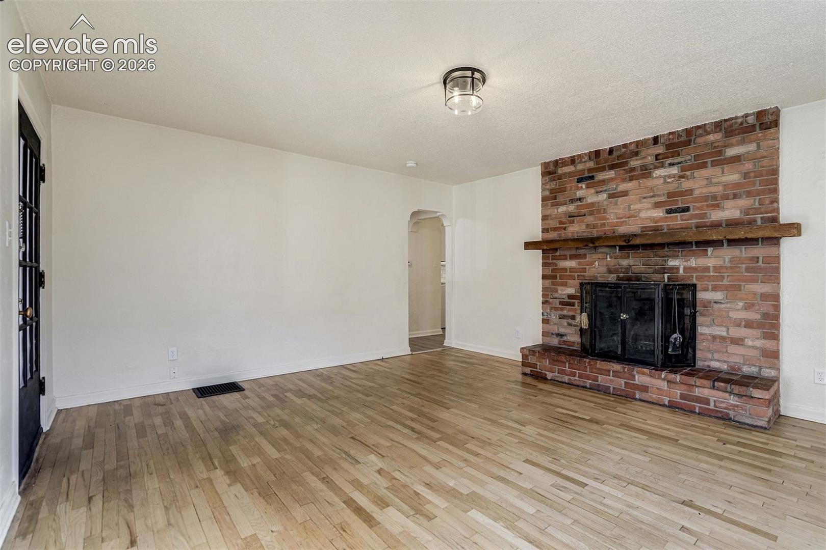 Living area featuring wood-finish flooring, a floor-to-ceiling brick fireplace with a raised hearth, a prominent wood mantel, an arched doorway, and a multi-pane glass door