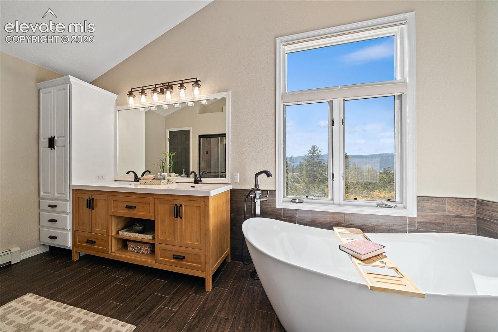Freestanding soaking tub with a floor-mounted faucet, wood-finish vanity with dual sinks, built-in floor-to-ceiling cabinetry, dark tile flooring, and a large picture window