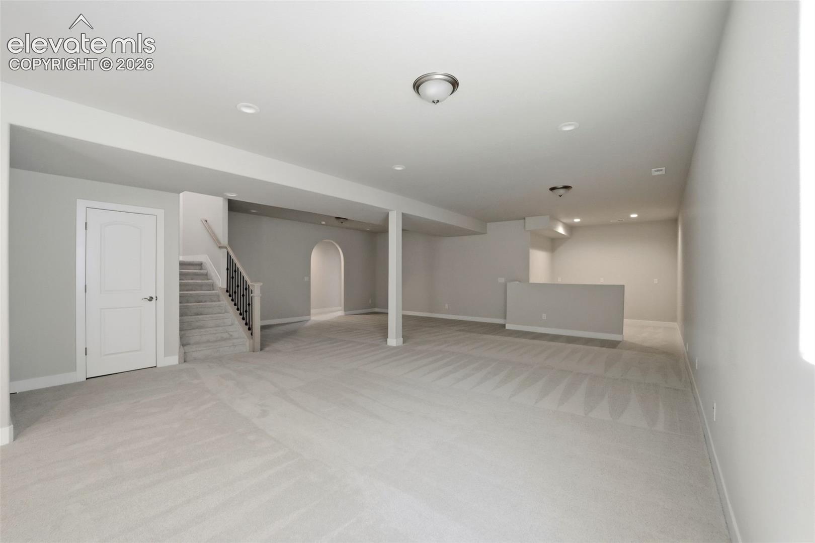 Open-concept room featuring wall-to-wall carpeting, recessed lighting, a built-in partition wall, an arched entryway, and a staircase with a decorative metal baluster railing