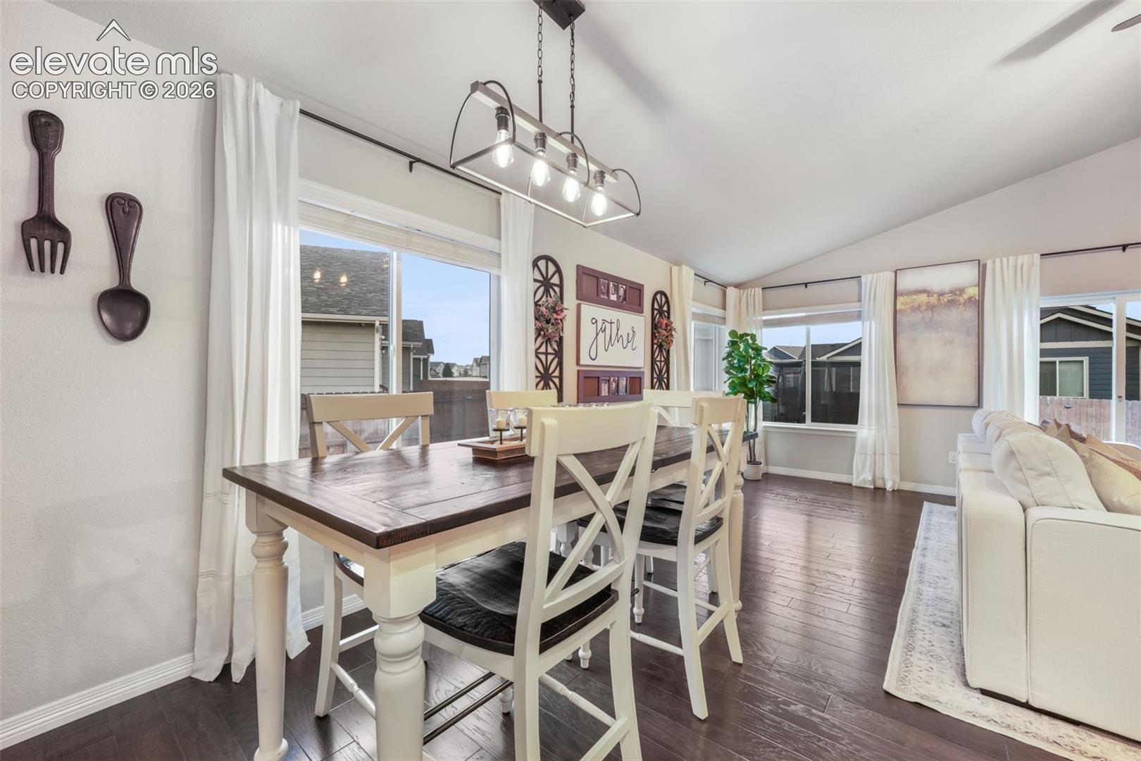 Vaulted ceiling with a linear chandelier, large windows with curtain rods, dark wood-finish flooring, and white wall paint