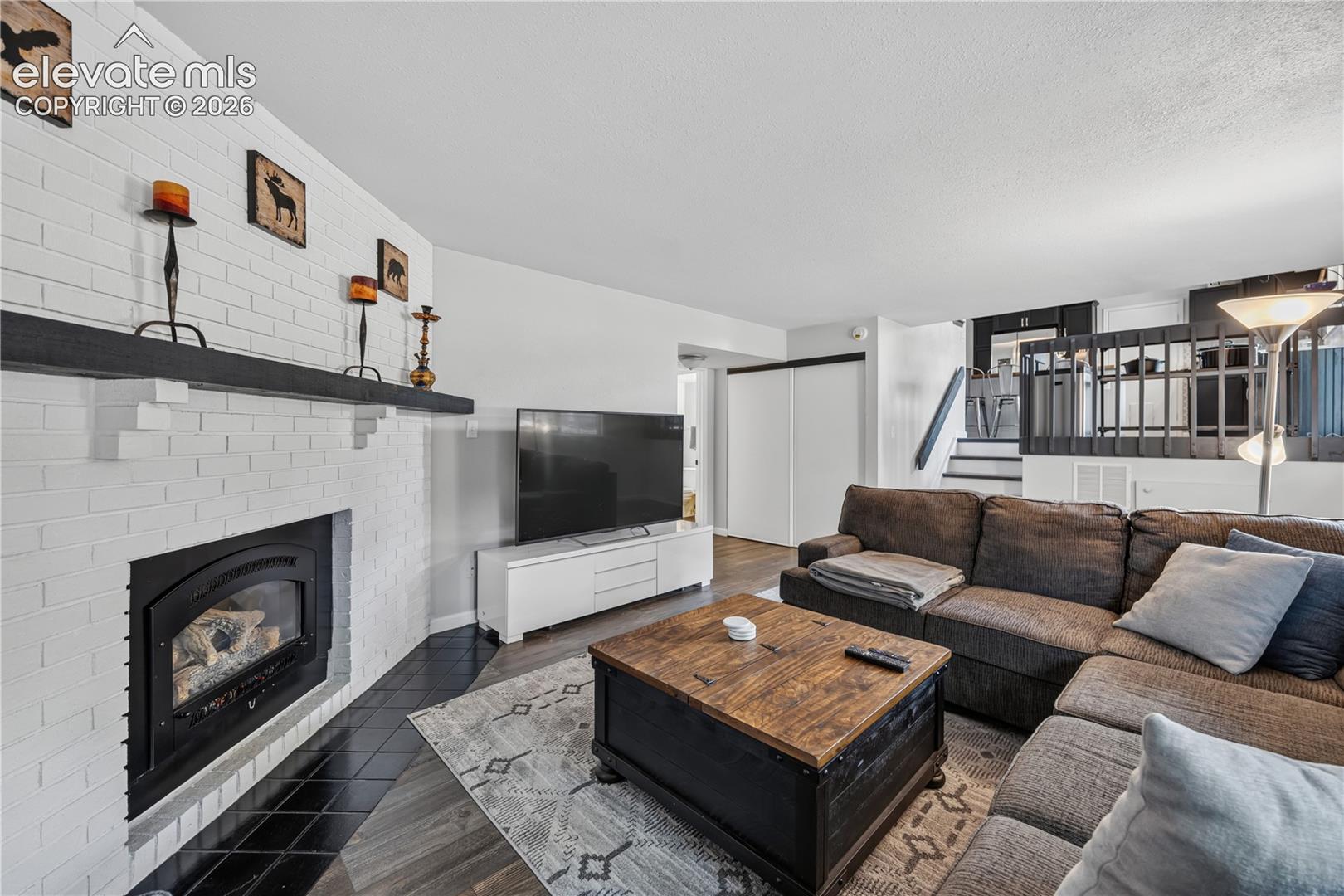 Living area featuring a white-painted brick fireplace with a dark mantle, black tile hearth, wood-finish flooring, and a decorative interior staircase railing