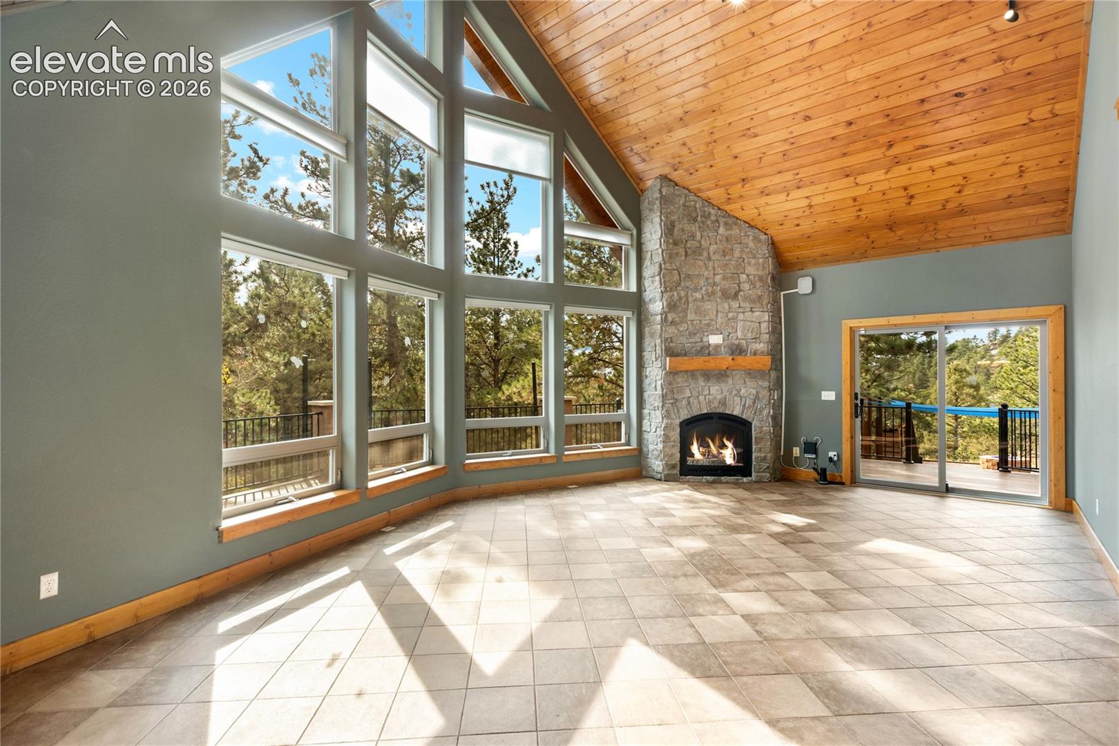 Vaulted wood-plank ceiling, floor-to-ceiling windows, stone-surround fireplace with a timber mantel, tile flooring, and sliding glass door access to the exterior