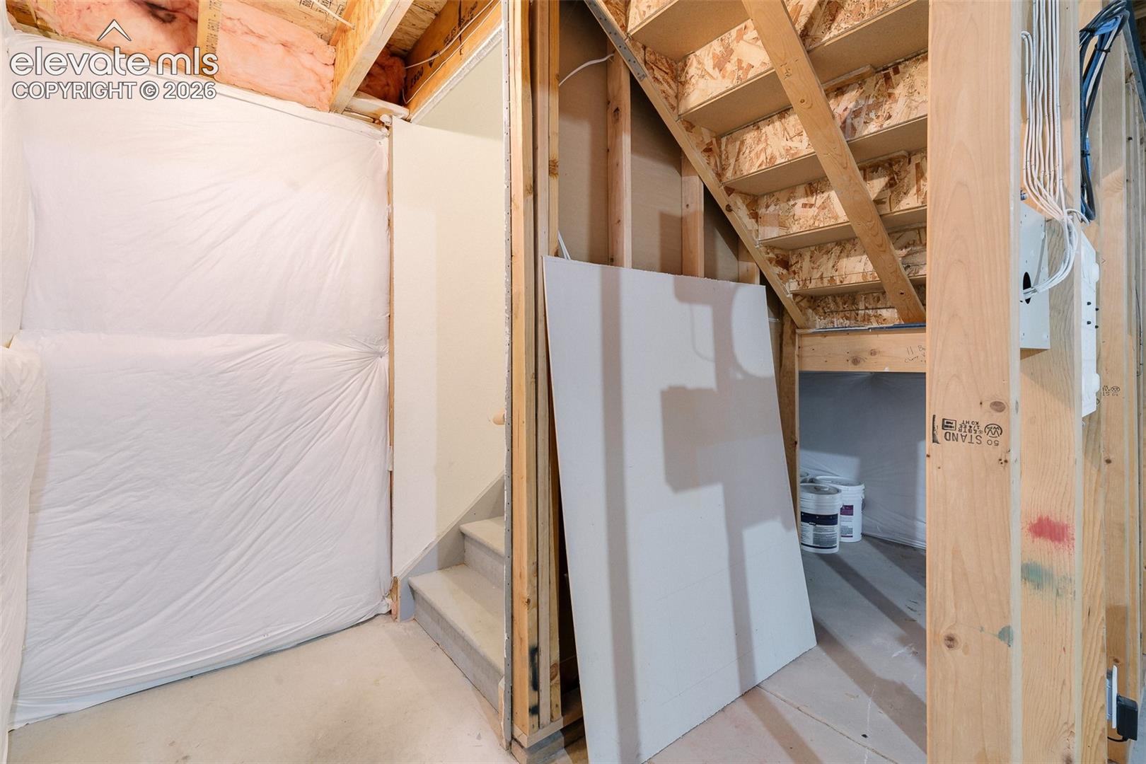 Unfinished lower level featuring exposed wood framing, concrete flooring, plastic vapor barrier wall covering, and timber staircase construction