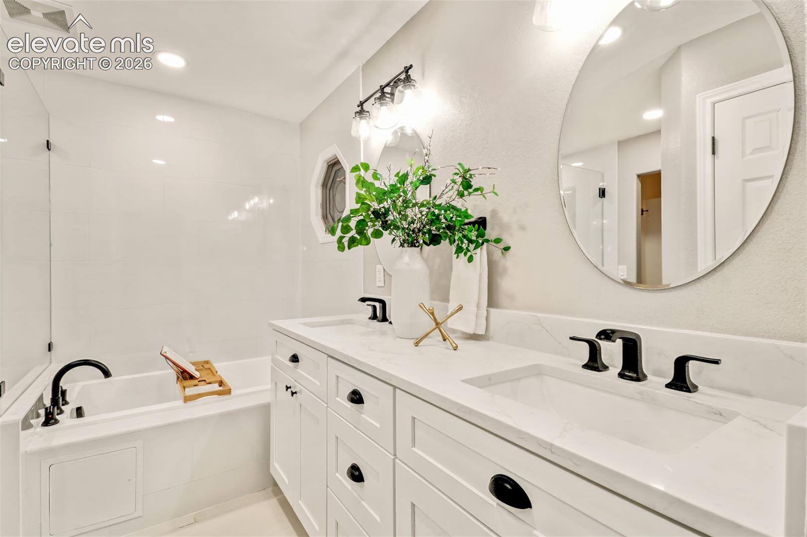Dual-sink vanity with stone-finish countertop, undermount basins, matte black widespread faucets, white Shaker-style cabinetry, and an integrated soaking tub with a glass panel