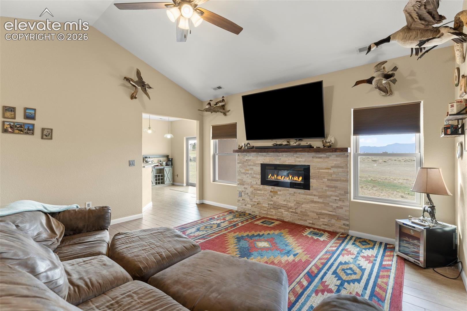 Vaulted ceiling, floor-to-ceiling stone-veneer fireplace with built-in electric insert, wood-finish flooring, and expansive windows