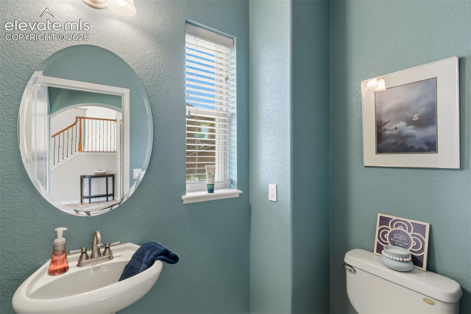 Pedestal sink with a polished chrome faucet, an oval frameless mirror, and a window with horizontal blinds