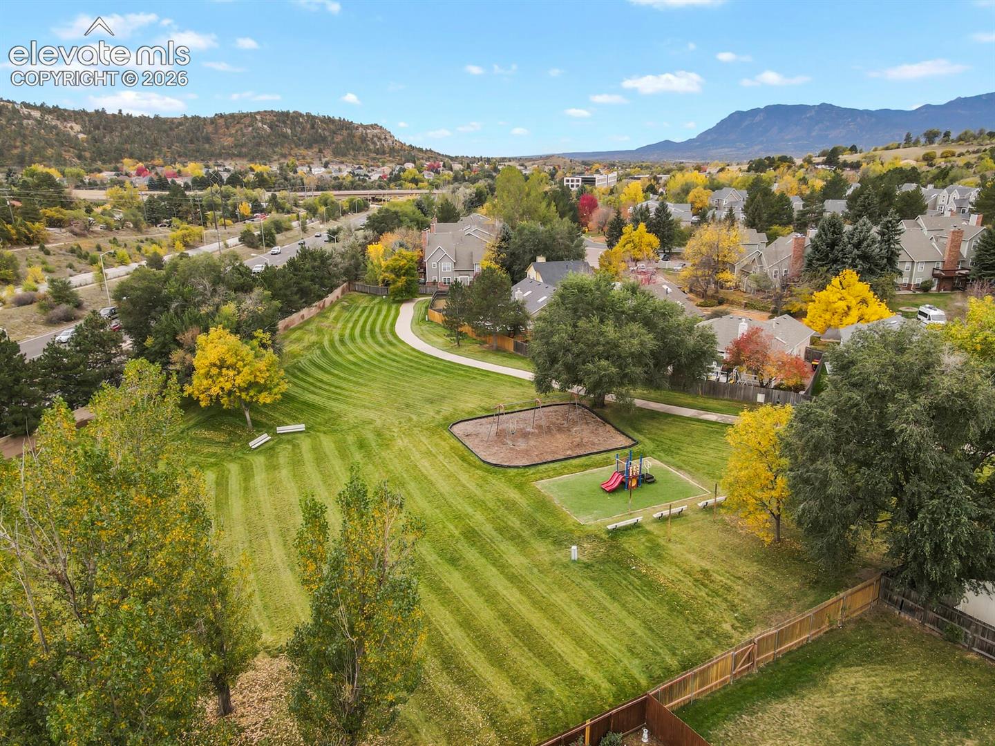 Expansive landscaped green space featuring mature trees, a paved walking path, playground equipment, and mountain backdrop