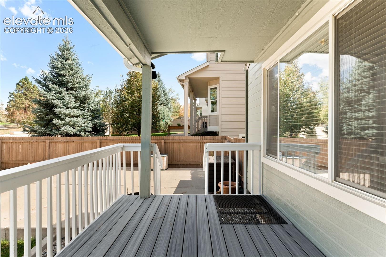 Covered deck featuring gray wood-finish planks, white baluster railings, and stairs leading to a concrete patio enclosed by a wood privacy fence
