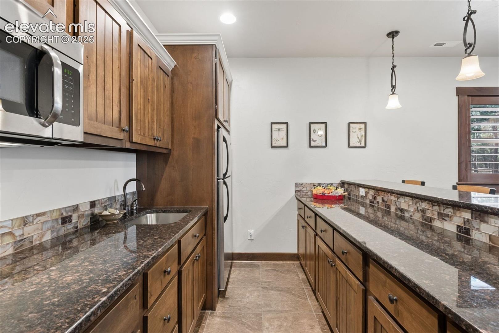 Kitchen featuring wood cabinetry, stone-finish countertops, decorative mosaic tile backsplash, built-in stainless steel appliances, and pendant lighting