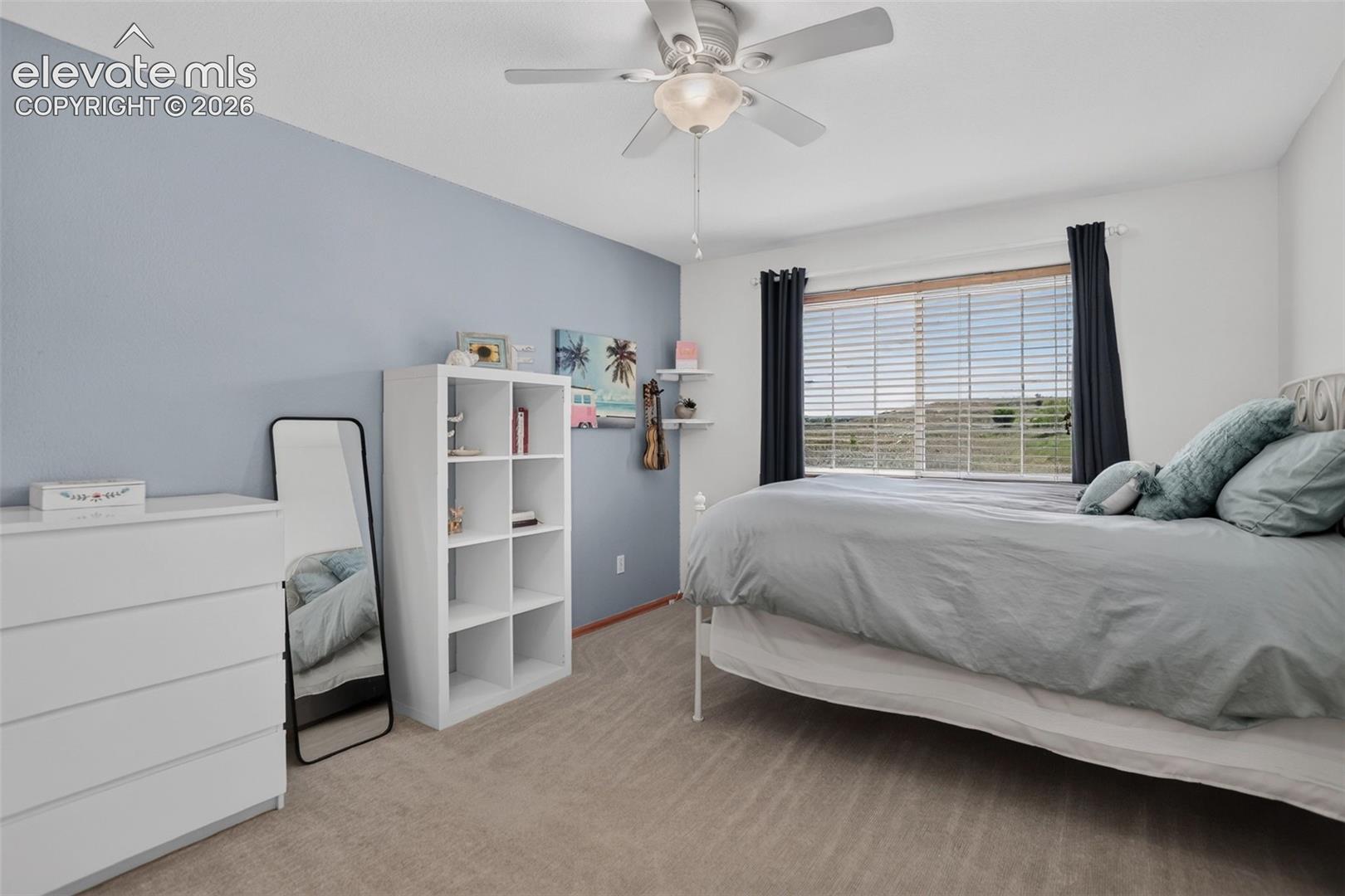 Carpeted bedroom featuring a light blue accent wall, a ceiling fan with integrated lighting, a window with horizontal blinds and dark curtains, and a built-in floating shelf