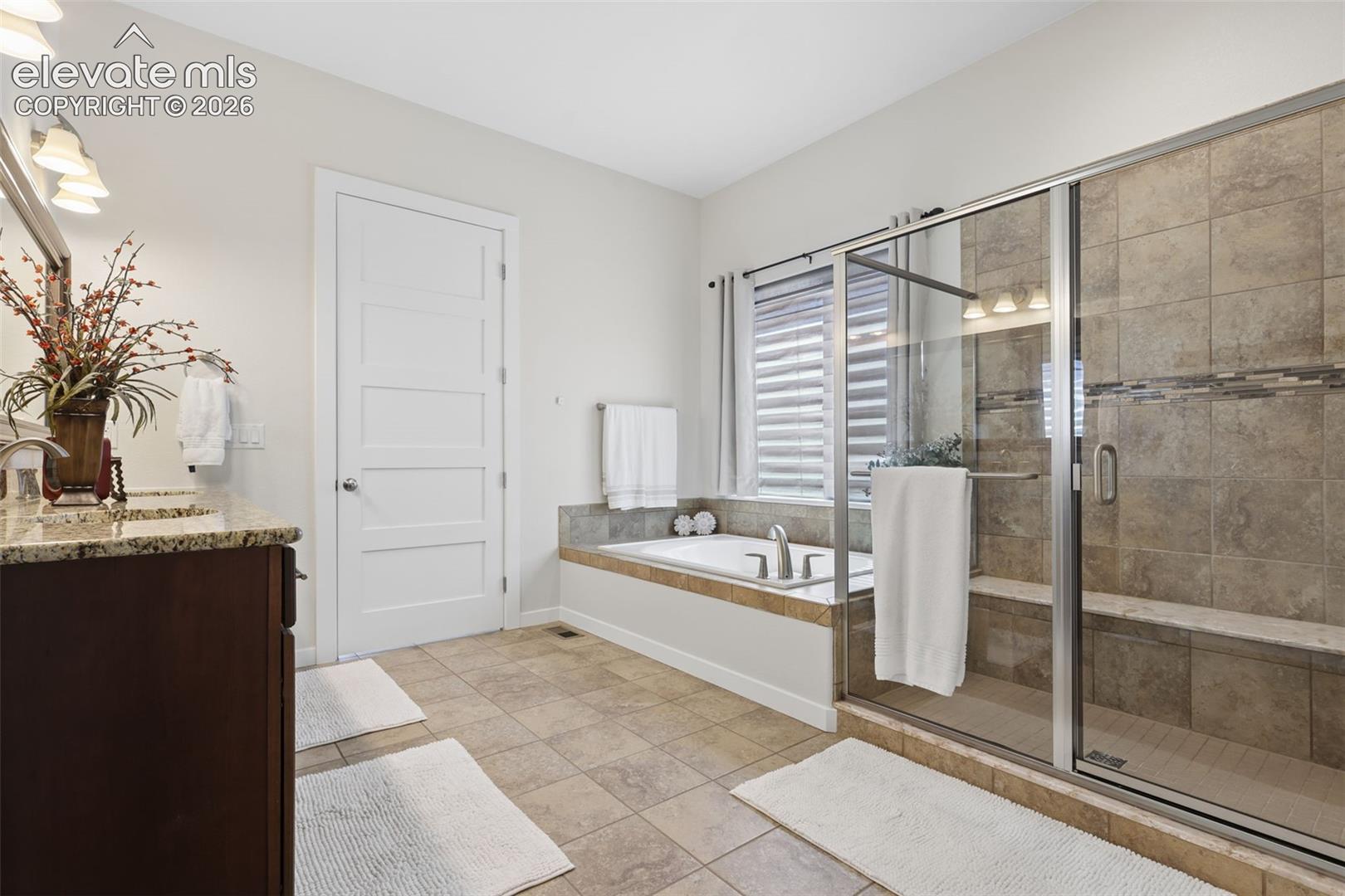 Dual sink vanity with granite countertop, built-in soaking tub, tiled walk-in shower with bench, and tiled flooring