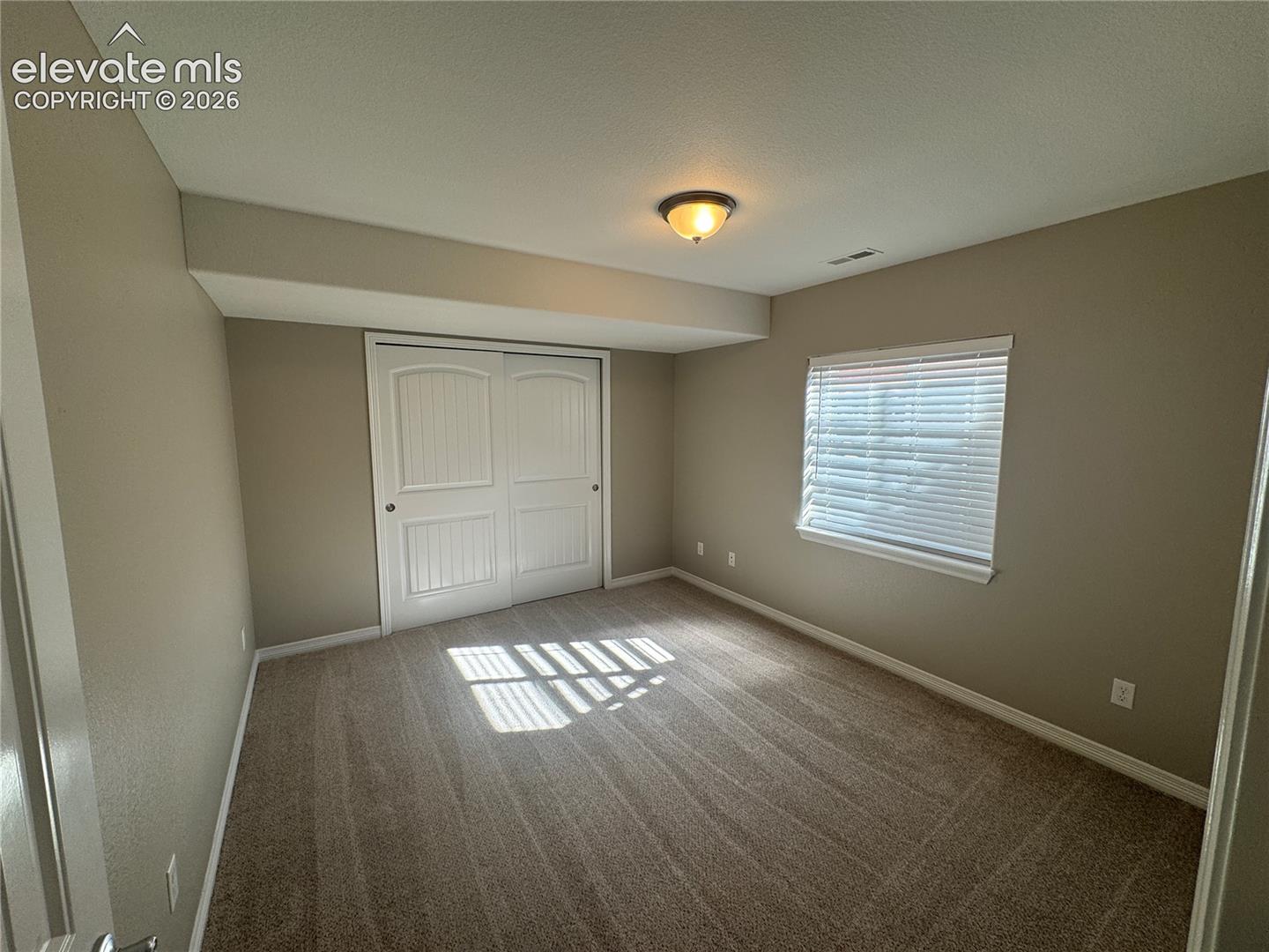 Neutral-toned bedroom featuring carpeting, ceiling-mounted light fixture, sliding panel closet doors, and window with horizontal blinds