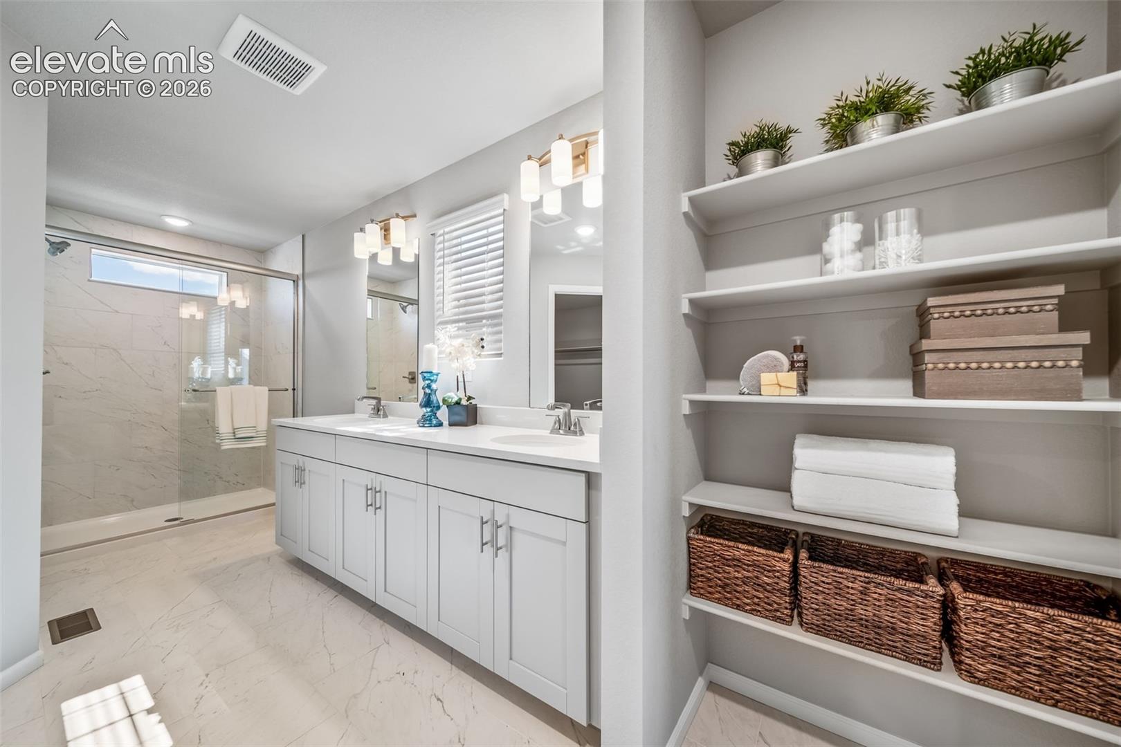 Dual vanity with light gray cabinetry, built-in shelving, glass-enclosed shower with a transom window, and tile flooring
*photos of a model home*