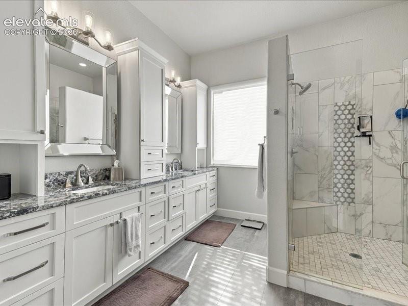 Dual vanity with granite countertops, extensive white cabinetry, large walk-in shower with a built-in bench, glass enclosure, and decorative tile accents, and light-toned floor tiling