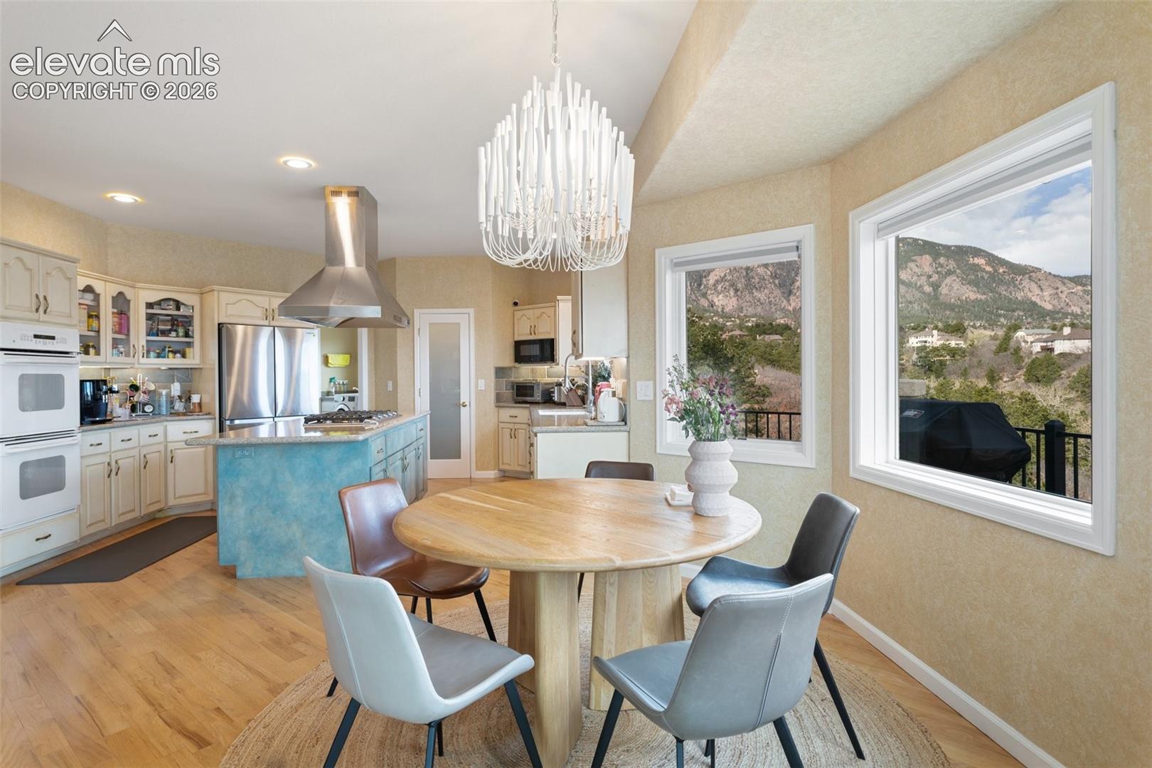Open-concept kitchen and dining area featuring light wood-finish flooring, a contemporary chandelier, two large windows, a stainless steel range hood, and a kitchen island with a cooktop