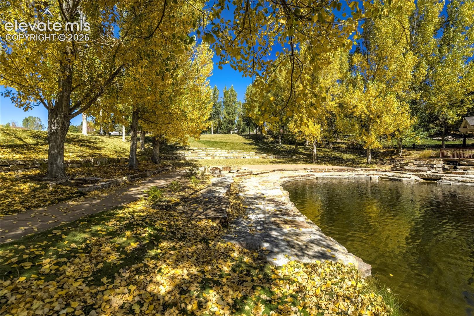 A peaceful neighborhood pond reflects the changing colors of the seasons, surrounded by mature trees + natural stone detailing that create a beautiful setting for a leisurely walk or quiet moment outdoors.