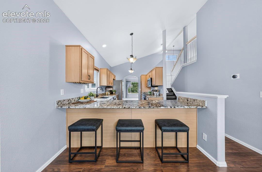 Kitchen featuring a breakfast bar with natural stone countertops, light wood-finish cabinetry, vaulted ceilings, and dark wood-finish flooring