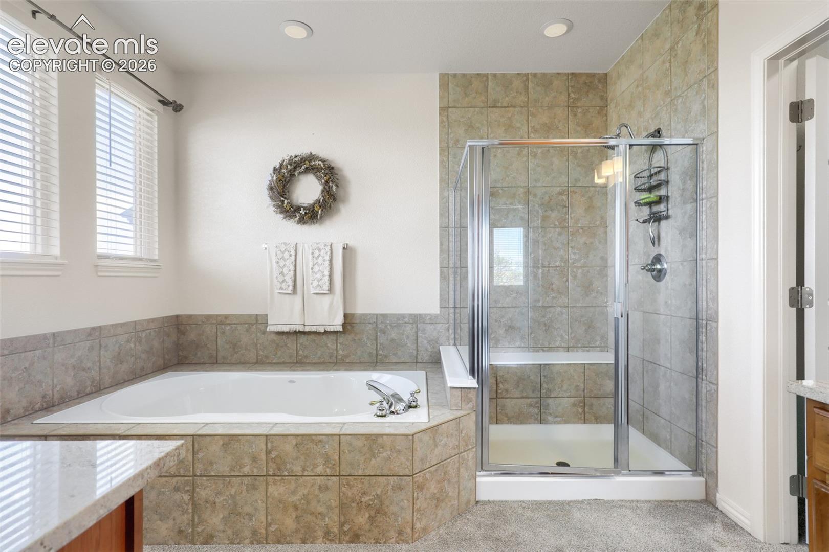 Designed for relaxation, this 5-piece primary bathroom features a deep drop-in soaking tub surrounded by custom stone-style tile and set beside bright windows with horizontal blinds. Directly adjacent, a spacious glass-enclosed shower boasts full tile surround and a convenient built-in bench seat.
